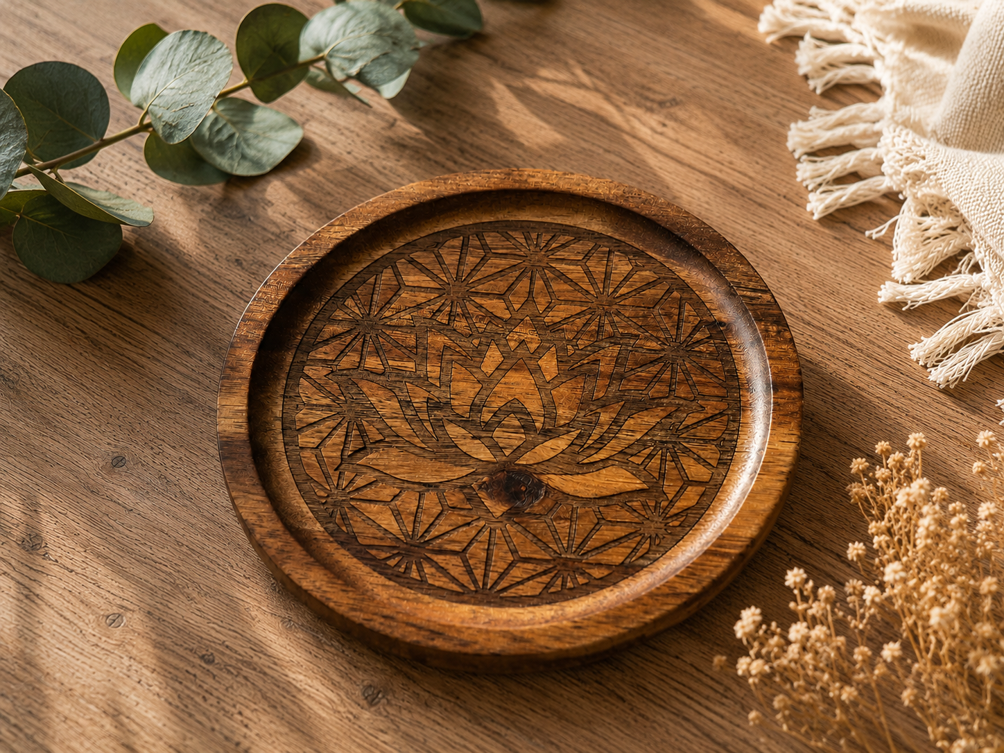 Plateau rond en bois gravé d'une fleur de lotus géométrique, posé sur une table en bois clair avec des fleurs séchées et une plante verte.
