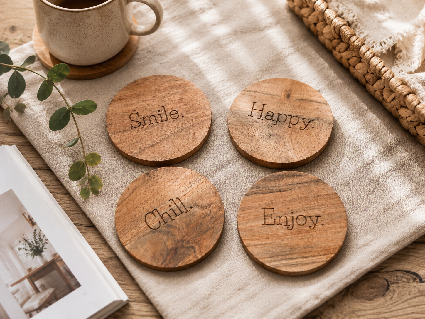 Collection de sous-verres en bois gravés Smile, Happy, Chill et Enjoy, mise en scène sur un tissu beige avec tasse et éléments décoratifs naturels.