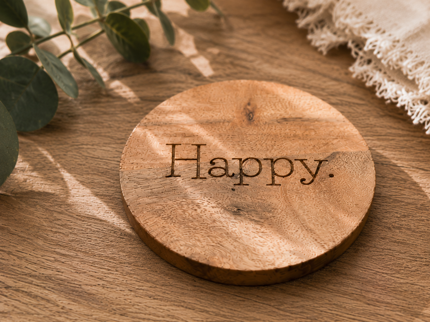 Sous-verre rond en bois naturel gravé du mot "Happy", photographié dans une ambiance chaleureuse aux tons naturels.